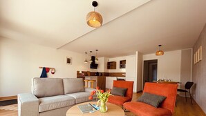 A modern living room with a grey sofa, a wooden coffee table, and orange armchairs. A kitchen is visible in the background with a microwave and wooden cabinets. - Ground floor holiday apartment for 4 people (Saint-Pierre-Quiberon)