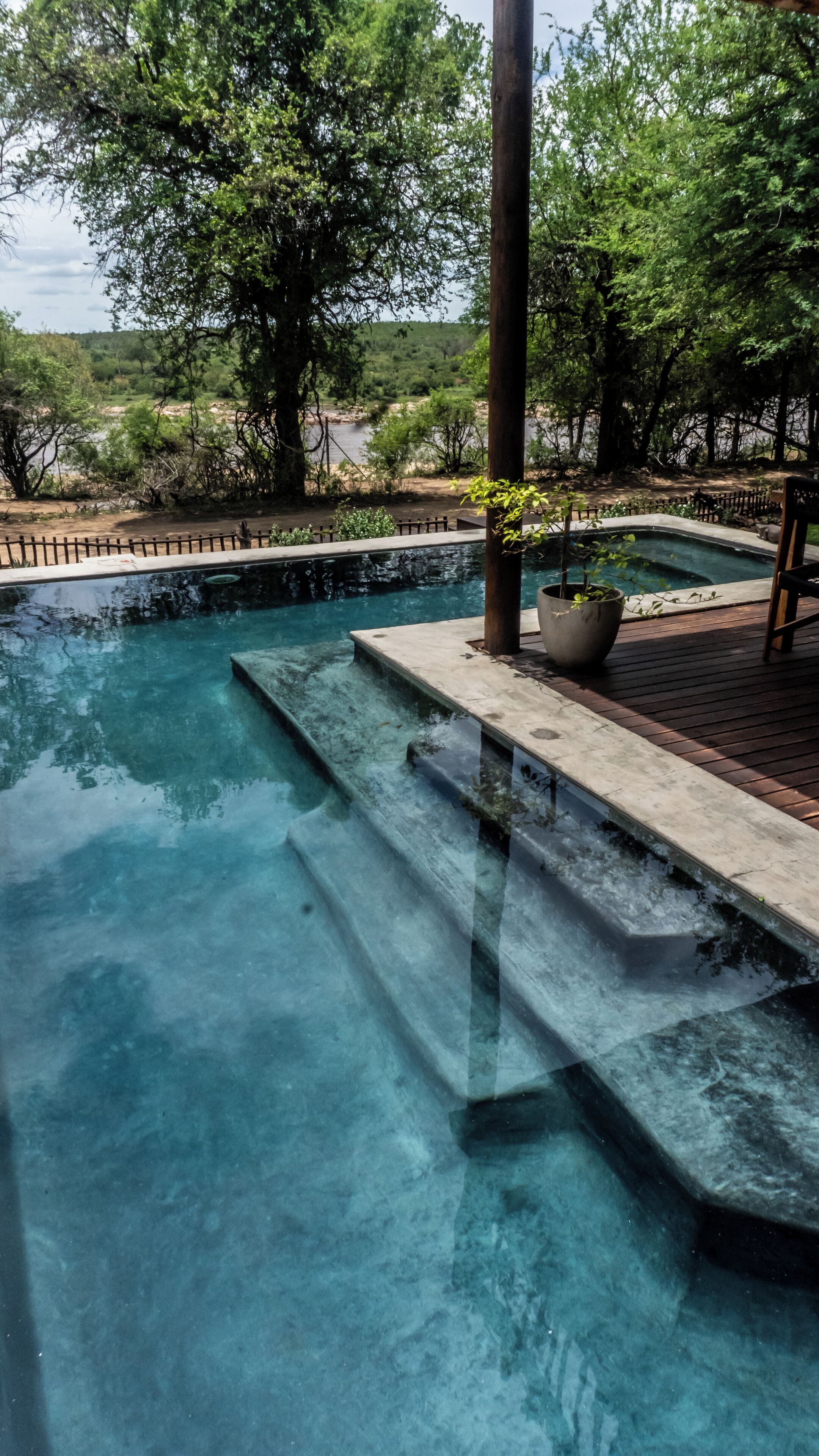 Une piscine aux eaux cristallines, accessible par des marches, entourée d’arbres et bordée d’une terrasse en bois.