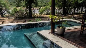 Une piscine aux eaux cristallines, accessible par des marches, entourée d’arbres et bordée d’une terrasse en bois.