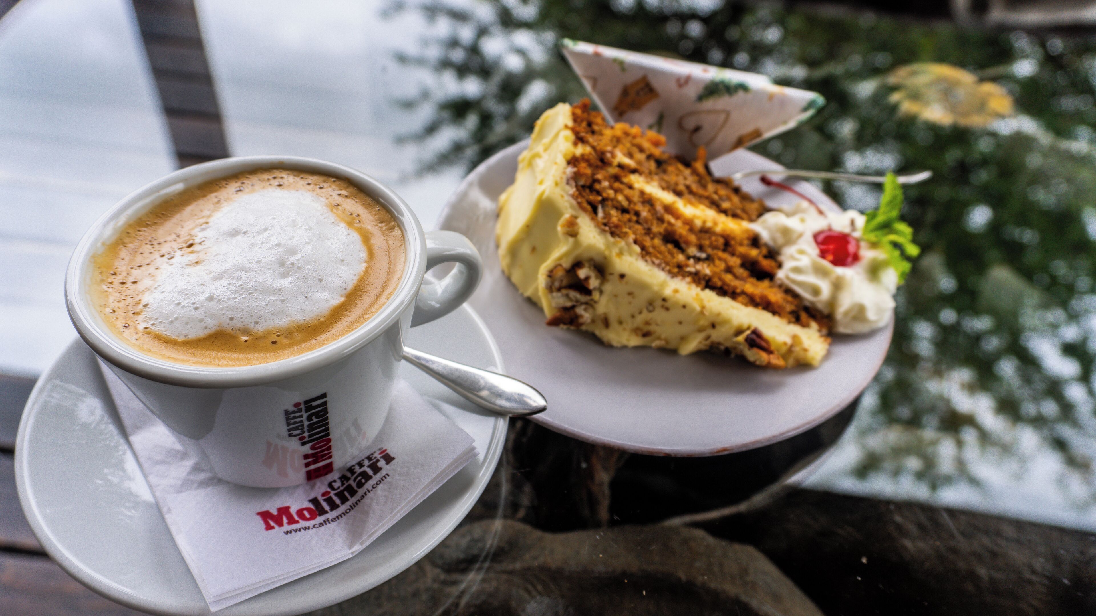 Uma chávena de café com espuma, uma fatia de bolo com chantilly e uma cereja, e uma colher num pires.