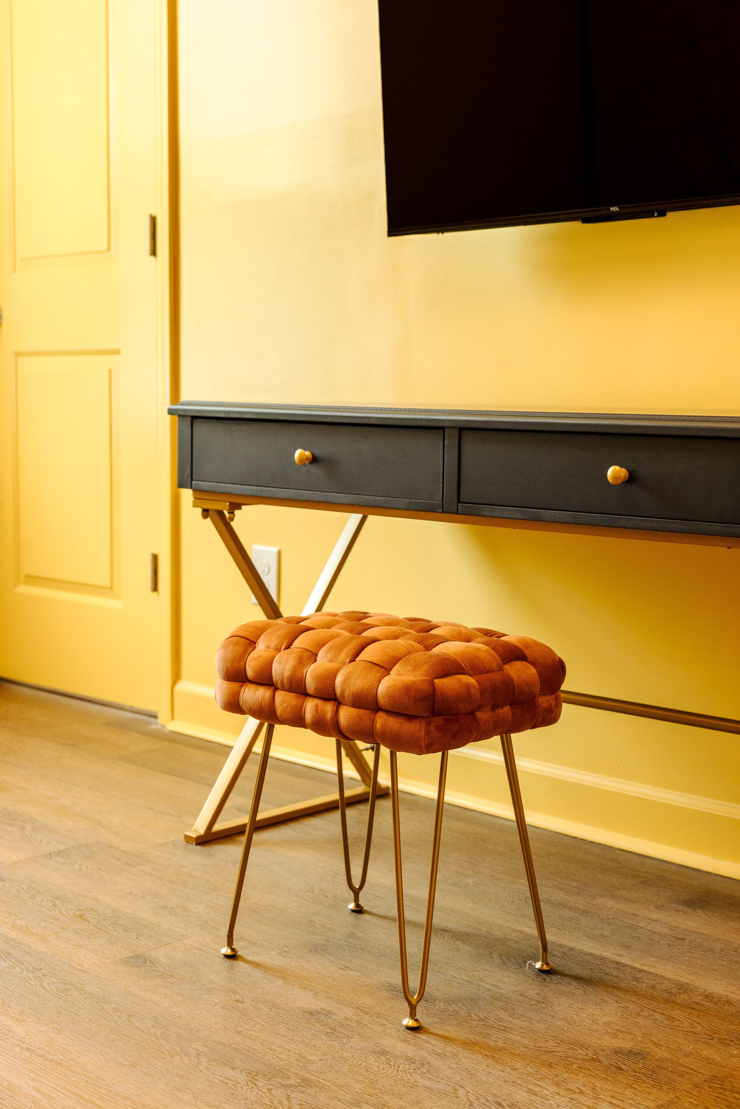 Una habitación con piso de madera, pared amarilla, escritorio negro con cajones y taburete tapizado marrón con patas metálicas.