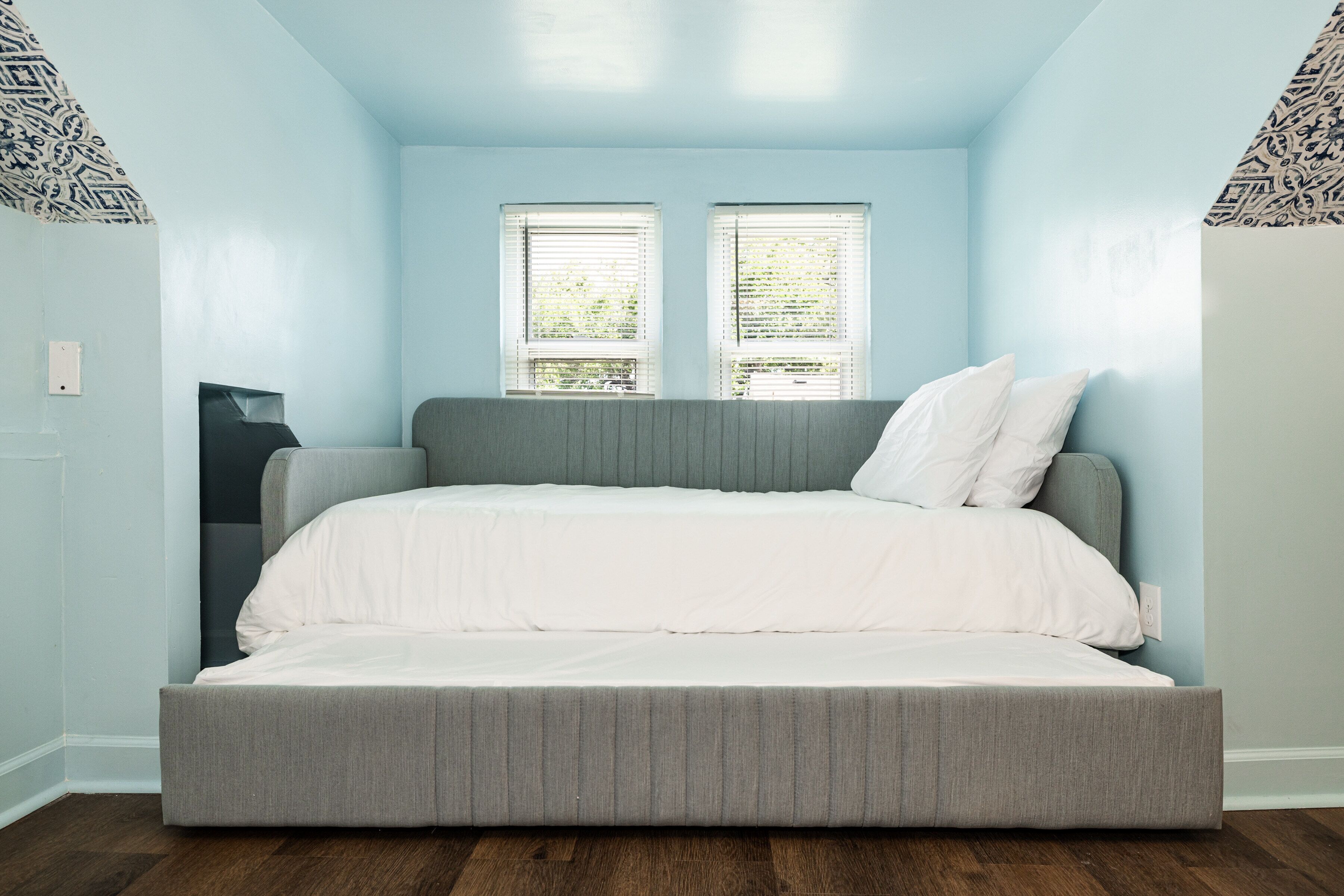 A bedroom with a grey bed, white bedding, two windows with blinds, and patterned wallpaper on the walls.