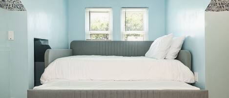 A bedroom with a grey bed, white bedding, two windows with blinds, and patterned wallpaper on the walls.