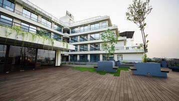 Un edificio moderno con varios pisos, grandes ventanales y una terraza de madera con macetas y asientos.