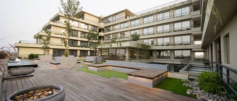 A modern multi-story building with a rooftop deck featuring wooden flooring, seating areas, and a water feature.