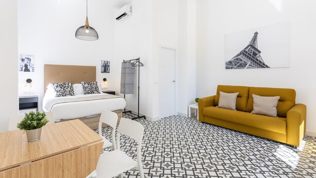 A modern bedroom with a bed, a yellow sofa, a dining table, and a patterned floor. - Holidays2Malaga Picassus Lofts (Málaga)