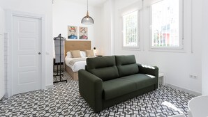 A modern bedroom with a green sofa, a bed with white bedding, a hanging light fixture, and a large window. - Holidays2Malaga Picassus Lofts (Málaga)