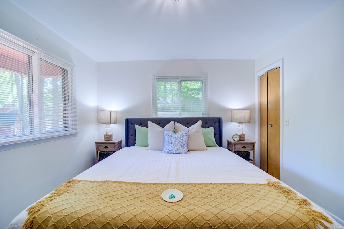 A neatly made bed with a patterned bedspread, two bedside tables with lamps, a window with blinds, and a door.
