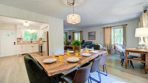 A spacious dining area with a wooden table set for six, surrounded by modern chairs. A chandelier hangs from the ceiling, and the room is well-lit with natural light from large windows.