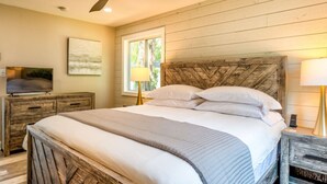 Een slaapkamer met een houten hoofdbord, een bed met witte beddengoed, een plafondventilator, een televisie op een houten dressoir, en een raam met natuurlijk licht.