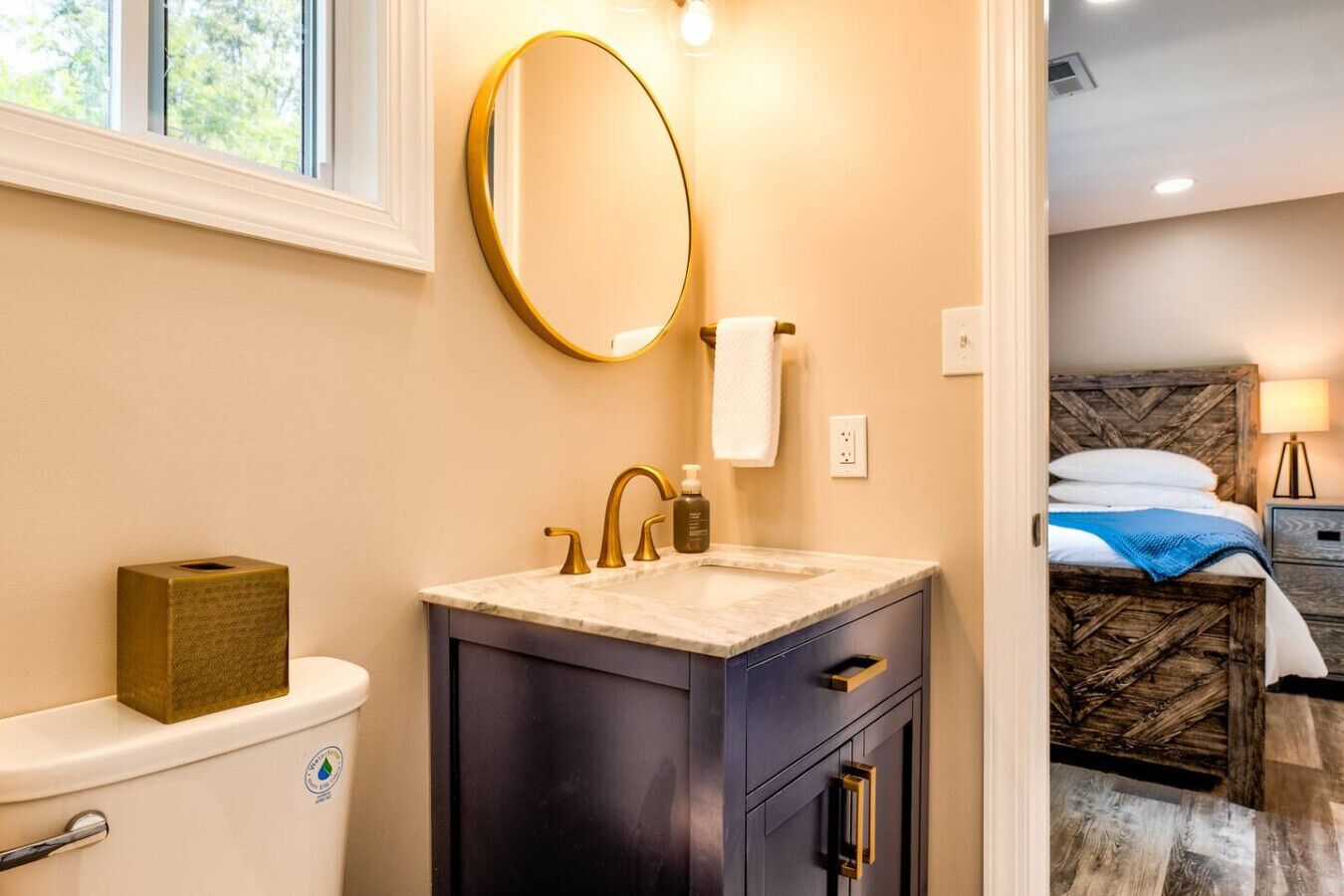 Une salle de bain avec des toilettes, un lavabo et un miroir. Une serviette est accrochée au mur et un flacon de savon est posé sur le lavabo. La pièce est dotée d’une tête de lit en bois et d’une table de chevet avec une lampe.