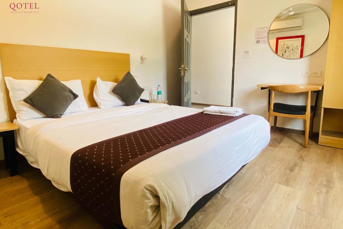 A hotel room with a large bed, two pillows, a wooden headboard, a bedside table with a bottle, a chair, a mirror, and a door.