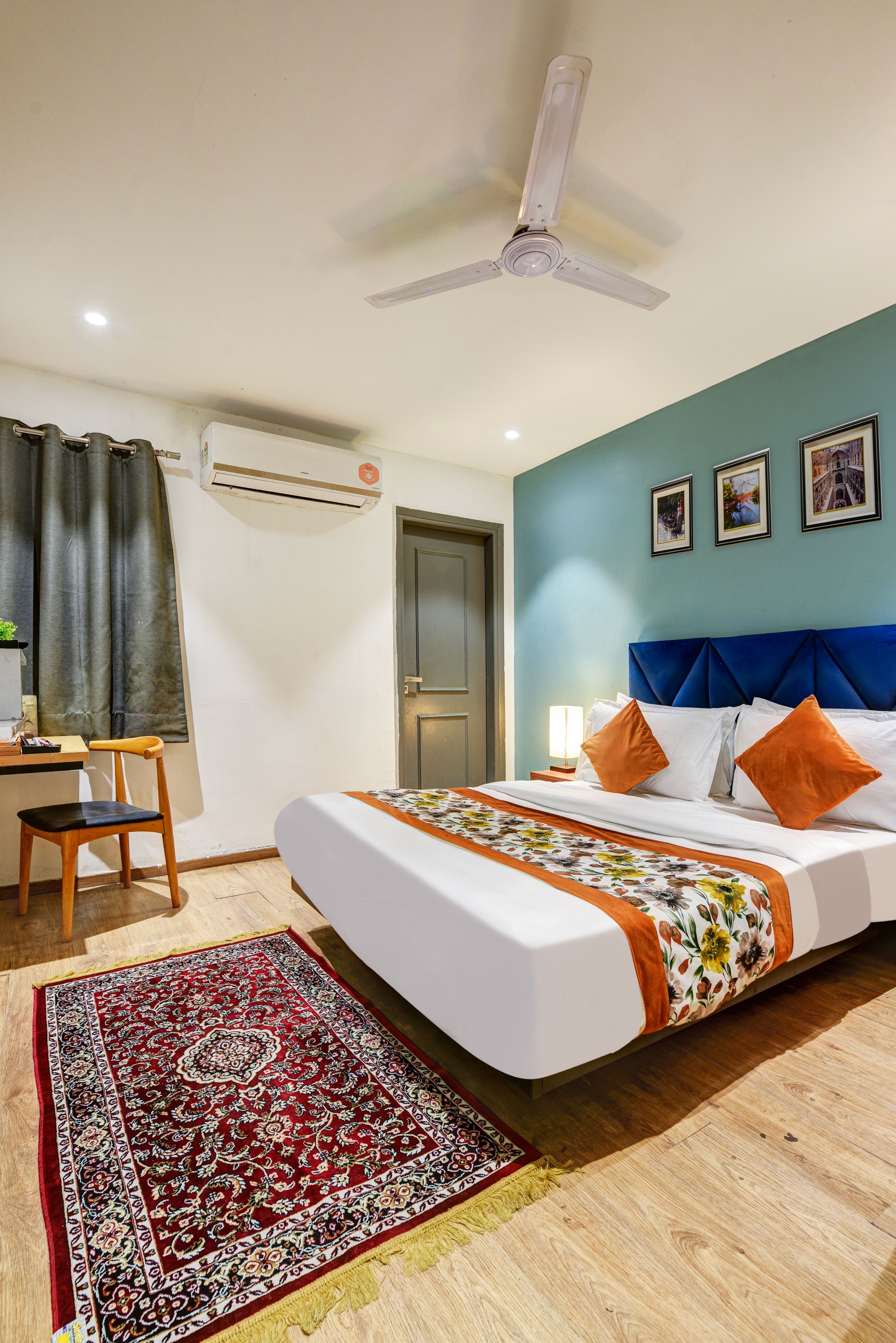 A hotel room with a bed, a desk, a chair, a ceiling fan, an air conditioning unit, a door, and a wall with three framed pictures.