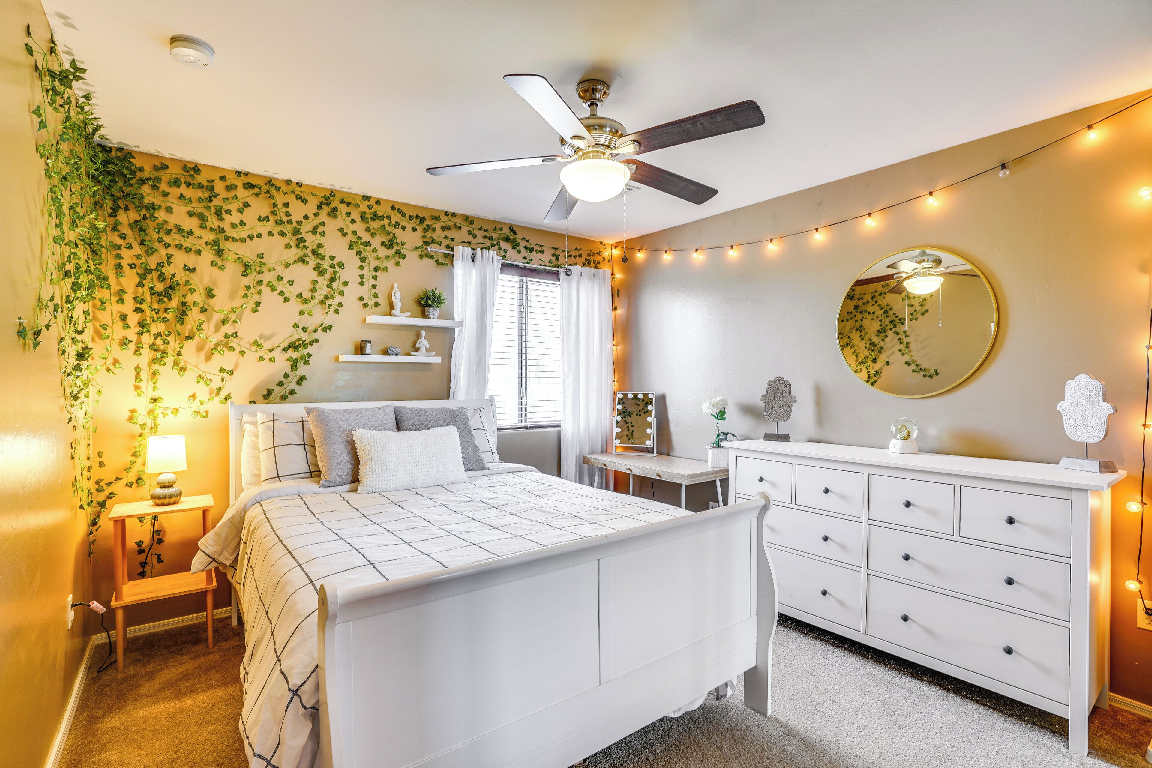 A bedroom with a bed, a dresser, a vanity, a ceiling fan, and string lights.