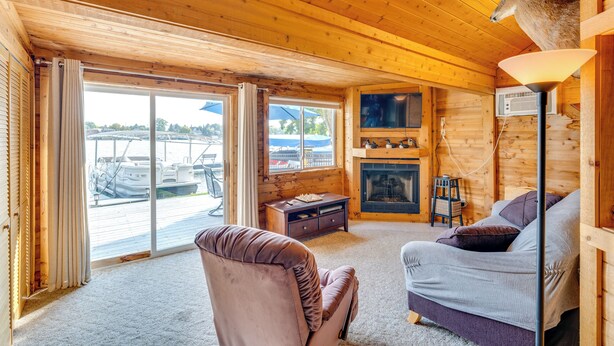 A cozy cabin interior with a fireplace, a plush sofa, and a large window overlooking a dock. - Lakefront Wisconsin Escape w/ Boat Dock & Kayaks! (Oconomowoc)