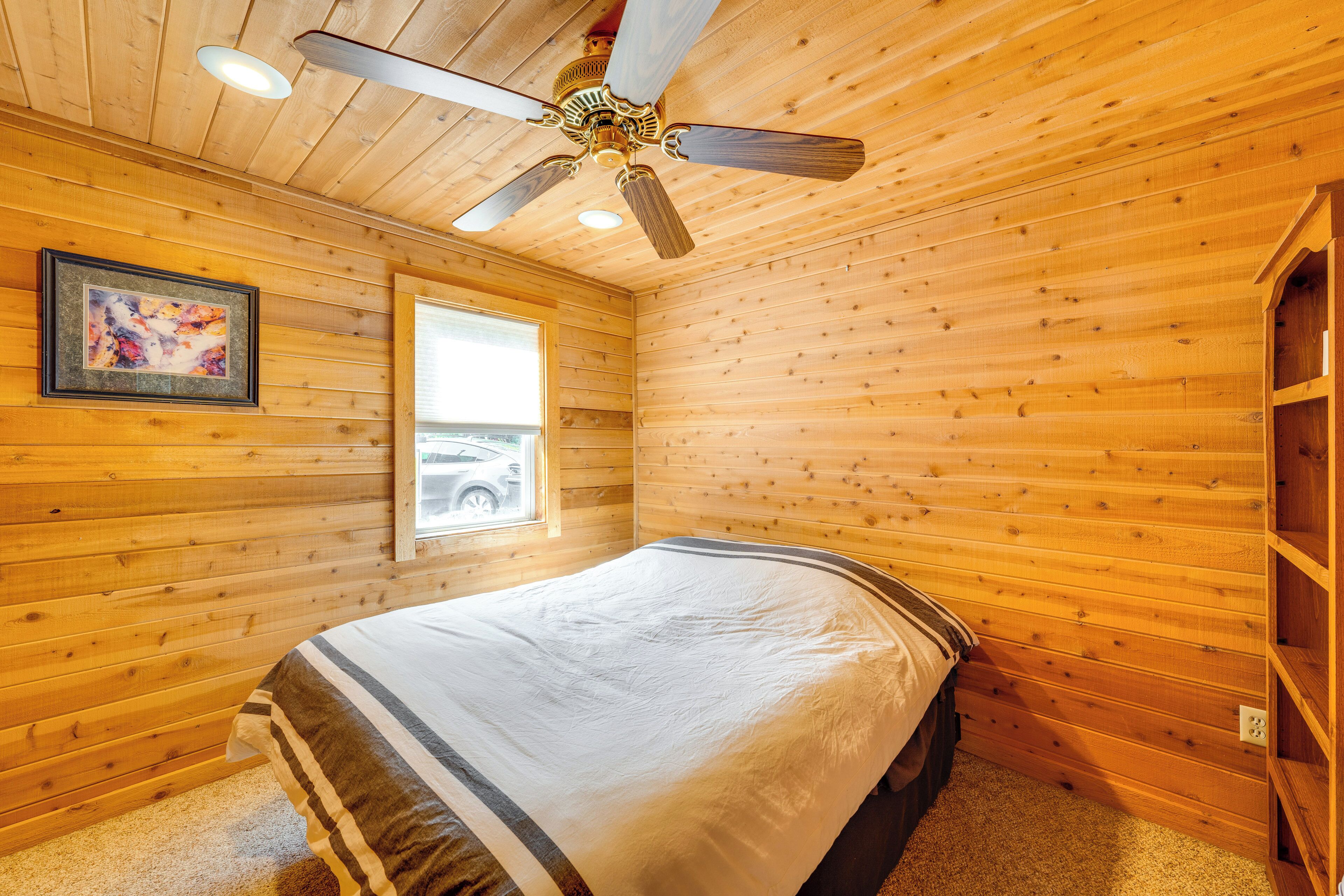 A wooden-paneled room with a bed, a ceiling fan, a window, and a framed picture on the wall.