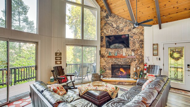 A spacious living room with a stone fireplace, leather sofa, and large windows overlooking a deck. - 2 Mi to Skypark at Santa's Village: Family Cabin (Lake Arrowhead)