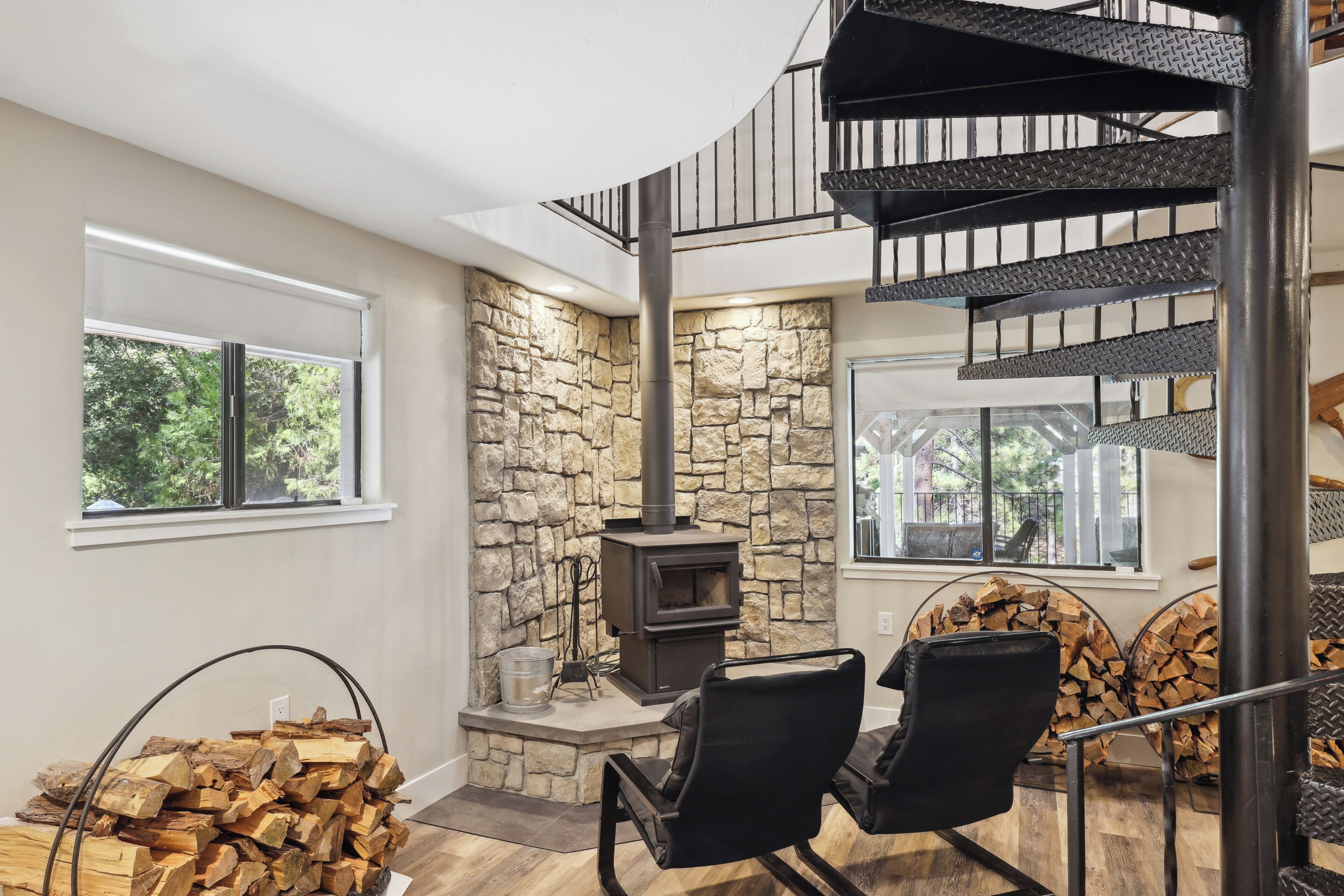 Un intérieur moderne avec un mur en pierre, un poêle à bois, un escalier en colimaçon et une vue sur l’extérieur.