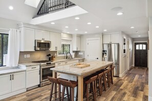 A modern kitchen with white cabinets, stainless steel appliances, a large island with stools, and a skylight. - Spacious Pine Mountain Lake Cabin Rental w/ Decks (Groveland)