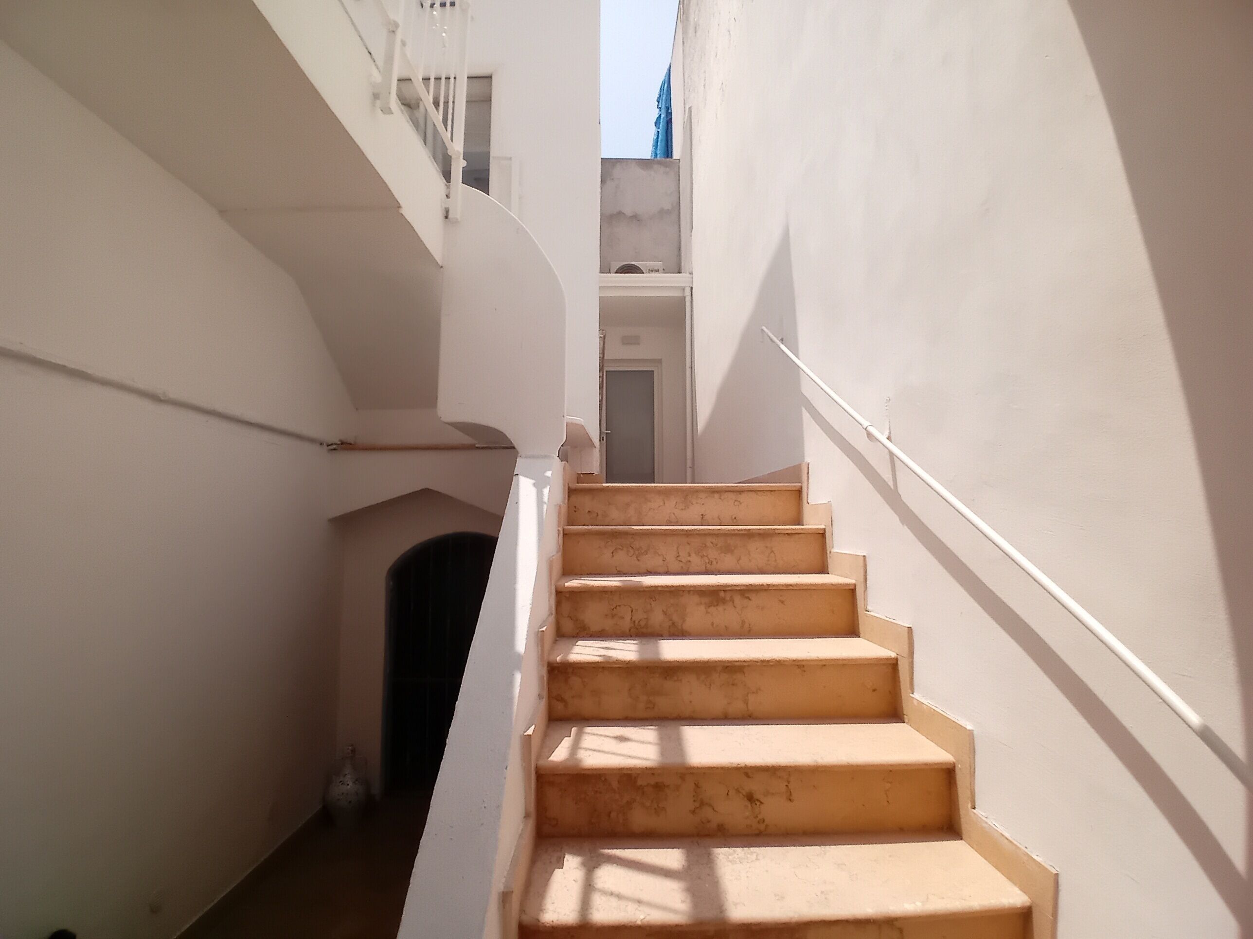 Una escalera estrecha de paredes blancas y barandilla de madera que lleva a una puerta en la parte superior.