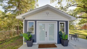 Ein kleines, eingeschossiges Haus mit einer weißen Tür und Fenstern, umgeben von einer Betonveranda und zwei großen Kübelpflanzen.