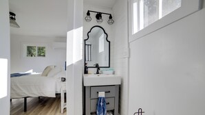 Ein modernes Badezimmer mit Waschbecken, Spiegel und Handtuchhalter. Im angrenzenden Raum befindet sich ein Bett mit weißer Bettwäsche.