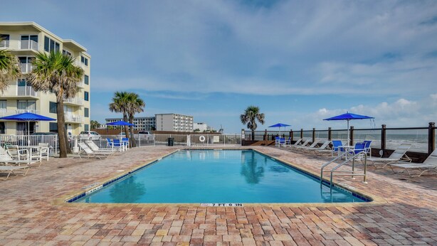 A swimming pool with lounge chairs and umbrellas, surrounded by a brick deck and a building with balconies. - Cozy Florida Getaway w/ Pool Access & Ocean Views! (Daytona Beach)