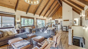 A spacious living area with a large sofa, a coffee table, and a dining table. There is a kitchen area visible in the background. - Cozy & Chic Lake Arrowhead Cabin w/ Deck! (Lake Arrowhead)