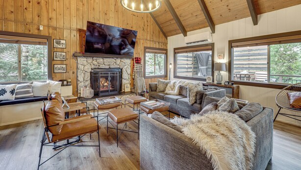 A spacious living room with a stone fireplace, leather seating, and large windows offering views of the outdoors. - Cozy & Chic Lake Arrowhead Cabin w/ Deck! (Lake Arrowhead)