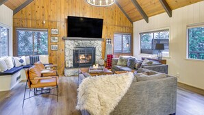 A cozy living room with a stone fireplace, wooden beams, a large TV, and comfortable seating. - Cozy & Chic Lake Arrowhead Cabin w/ Deck! (Lake Arrowhead)