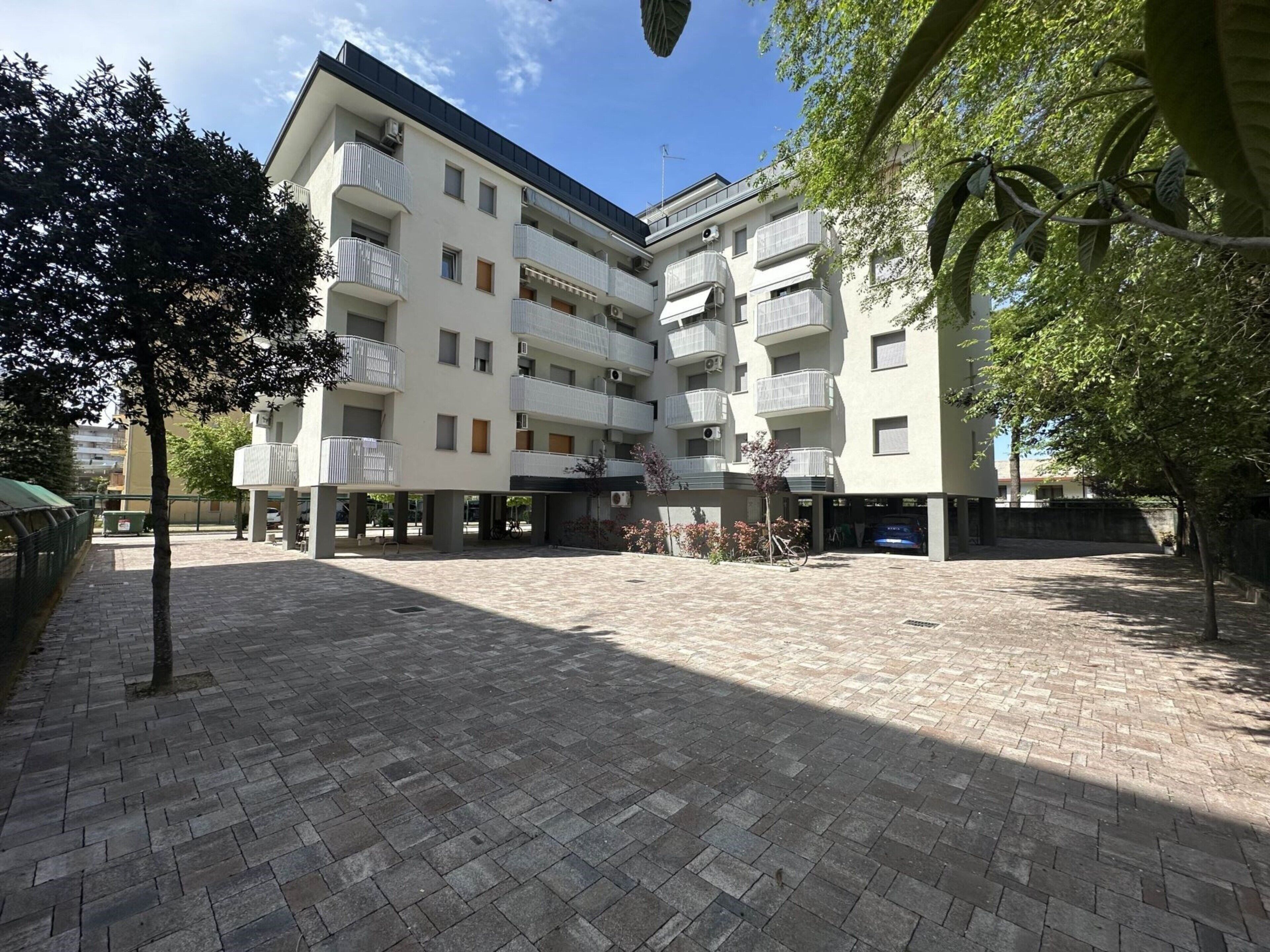 A multi-story residential building with balconies, a paved courtyard, and a bicycle parked in front.