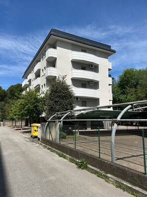 Un immeuble à plusieurs étages doté de balcons, d’un espace de stationnement clôturé et d’une poubelle jaune.