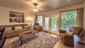 A living room with a brown sofa, a coffee table, a chair, and a ceiling fan. There is a large window with blinds and a view of greenery outside.