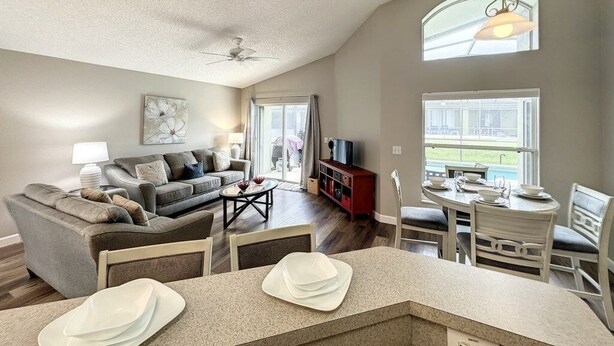 A spacious living room with a high ceiling, a ceiling fan, a dining area with a table and chairs, and a large window providing a view of the outdoors. - 4 Bed 3 Bath In A Gated Community With Pool 4 Bedroom Villa by RedAwning (Kissimmee)