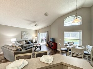 A spacious living room with a high ceiling, a ceiling fan, a dining area with a table and chairs, and a large window providing a view of the outdoors. - 4 Bed 3 Bath In A Gated Community With Pool 4 Bedroom Villa by RedAwning (Kissimmee)