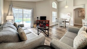 A spacious living room with a large sofa set, a coffee table, and a television. There is a dining area with a table and chairs, and a ceiling fan. - 4 Bed 3 Bath In A Gated Community With Pool 4 Bedroom Villa by RedAwning (Kissimmee)