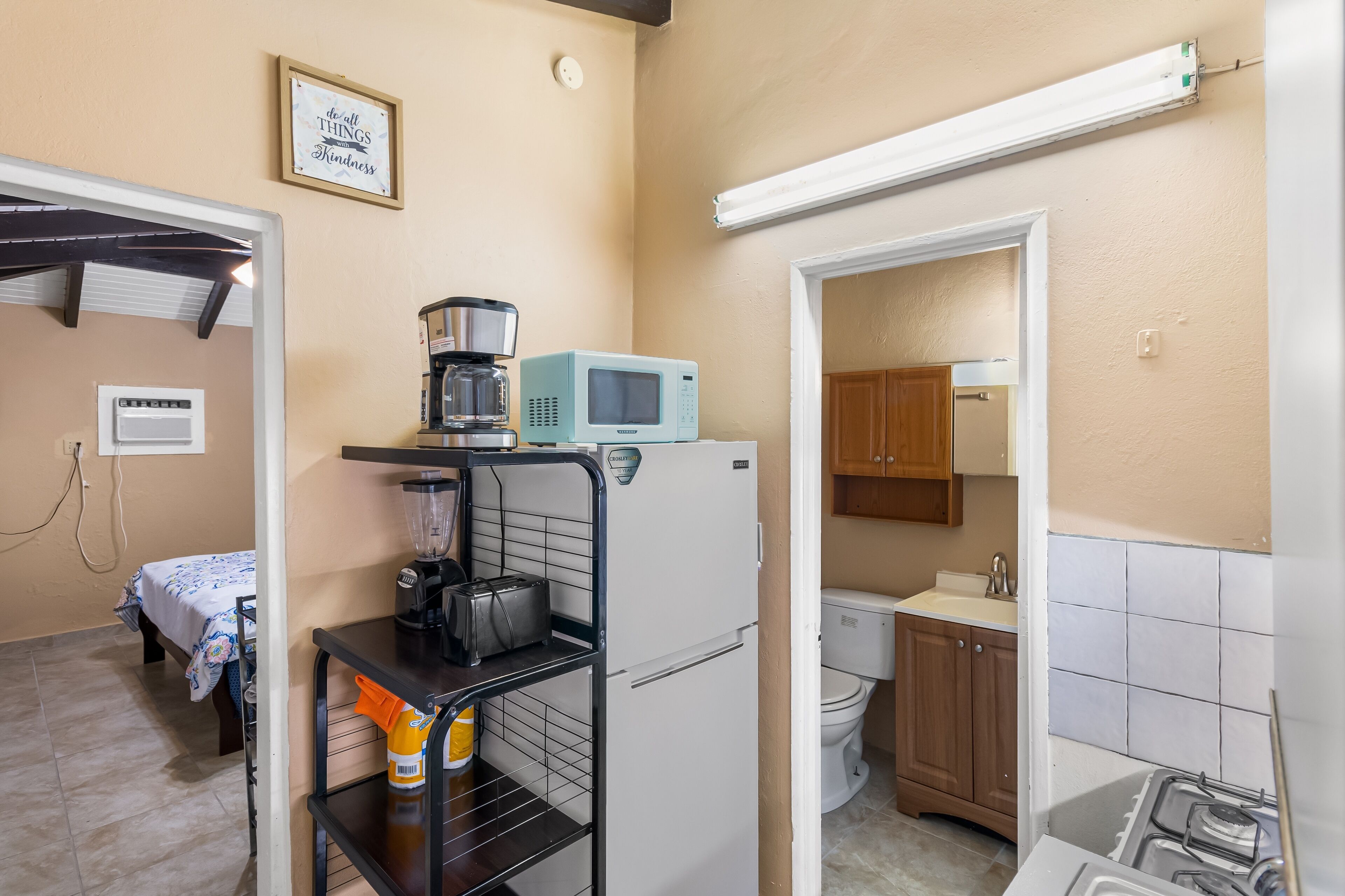 A compact kitchen with a microwave, coffee maker, and a small refrigerator. There is a bed with a white and blue patterned bedspread in the adjacent room.