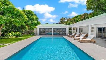A rectangular swimming pool with clear blue water, surrounded by a paved deck. There are lounge chairs with white cushions aligned along the pool's edge.