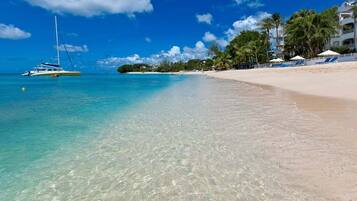 A beach with clear turquoise water, a sailboat, and a resort with palm trees and beach umbrellas.