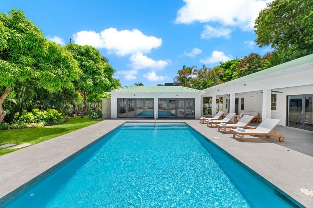 A rectangular swimming pool with clear blue water, surrounded by a paved deck. There are lounge chairs with white cushions aligned along the pool's edge.