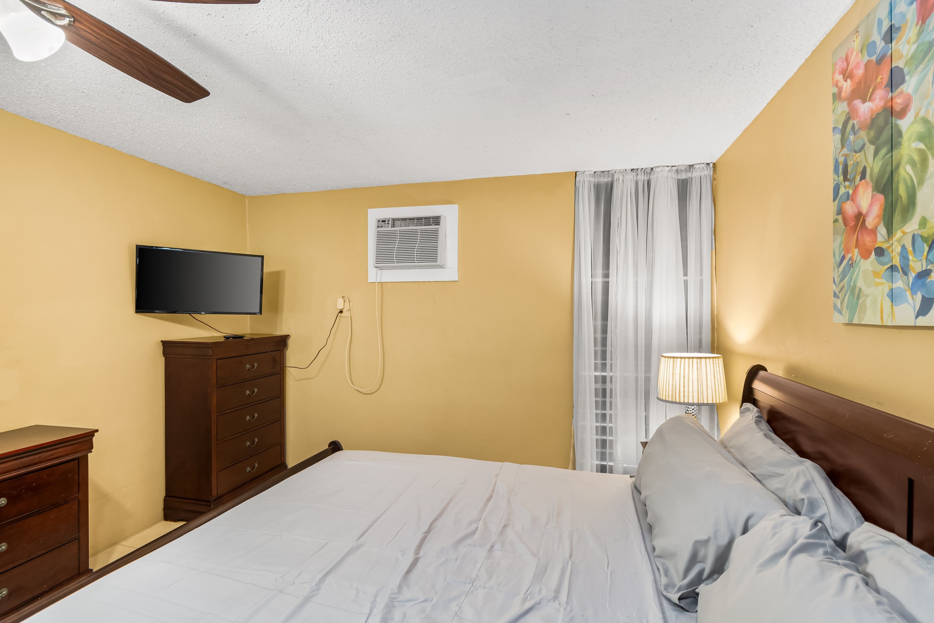 A bedroom with a bed, a TV, a dresser, a ceiling fan, a window with curtains, and a wall-mounted air conditioner.