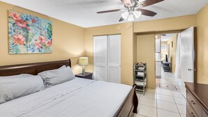 A bedroom with a bed, a ceiling fan, a nightstand with a lamp, a wardrobe, and a hallway leading to a bathroom.