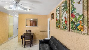 A living room with a black leather sofa, a dining table with chairs, a ceiling fan, and a wall with tropical artwork. - Yours For The Asking- Cozy, Caribbean, Condo 2 Bedroom Condo by RedAwning (Christiansted)