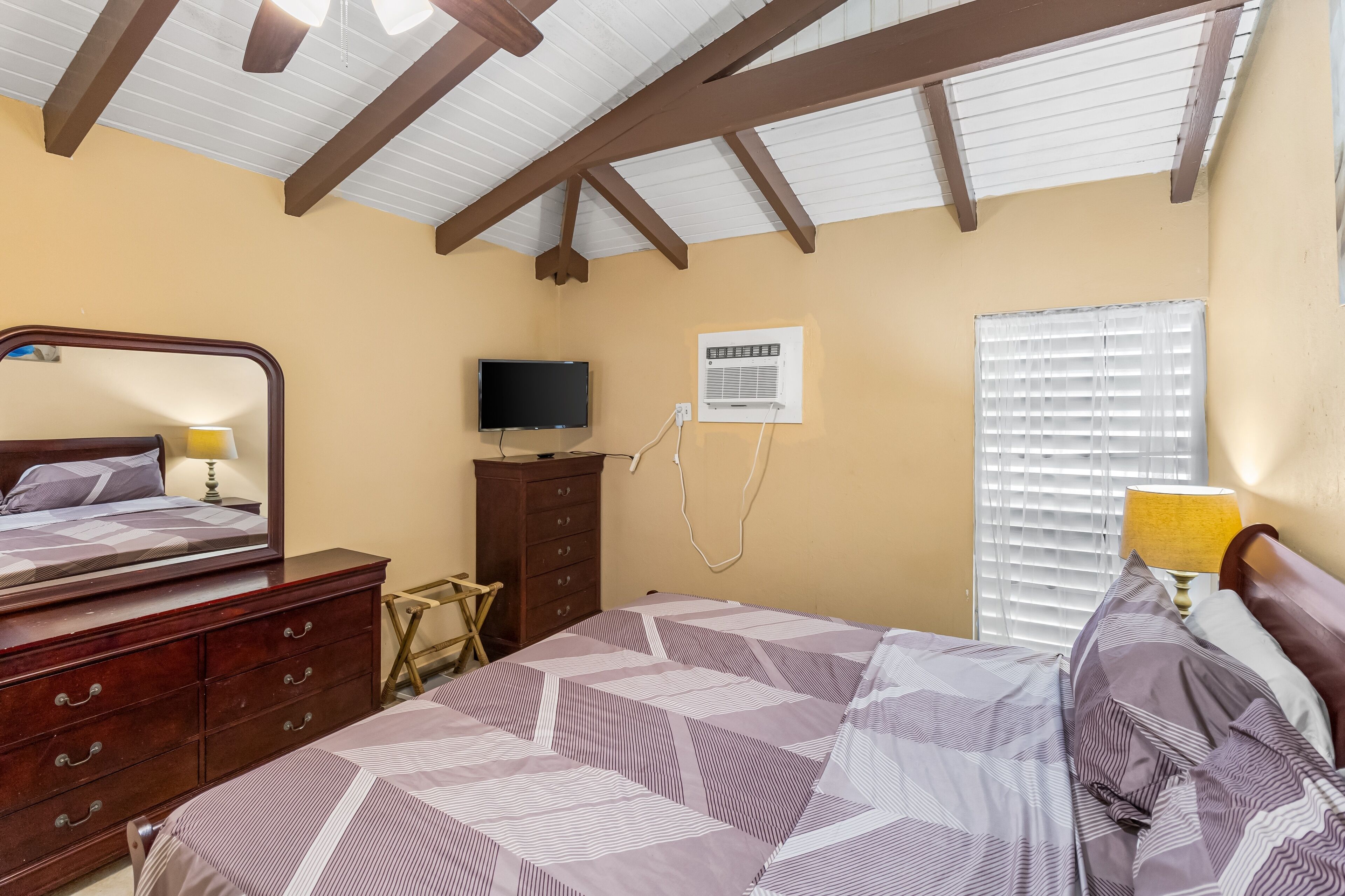 A bedroom with a bed, wooden dresser, TV, air conditioning unit, and window with blinds.