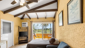 Une pièce comprenant un lit, un canapé, un balcon avec des chaises, un ventilateur de plafond et un tableau encadré au mur.