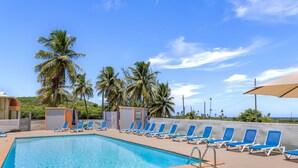 Une piscine bordée de chaises longues bleues, de palmiers et surplombée d’un ciel dégagé.