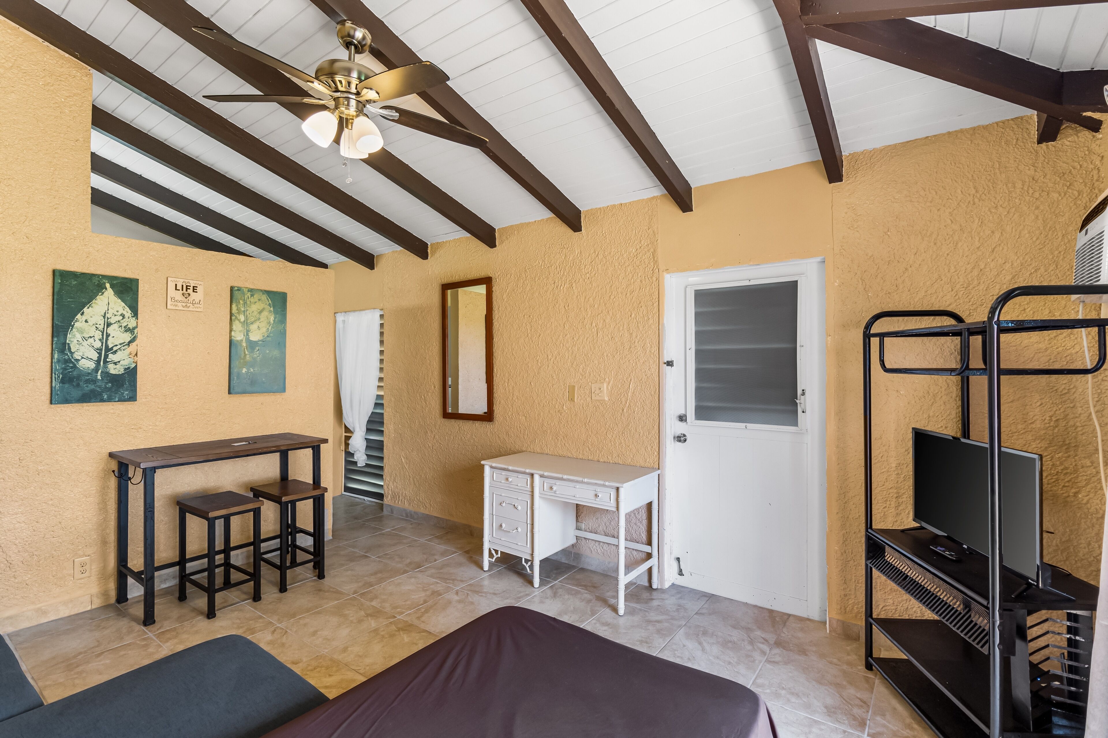 A room with a bed, a desk, a chair, a TV on a stand, and a ceiling fan.