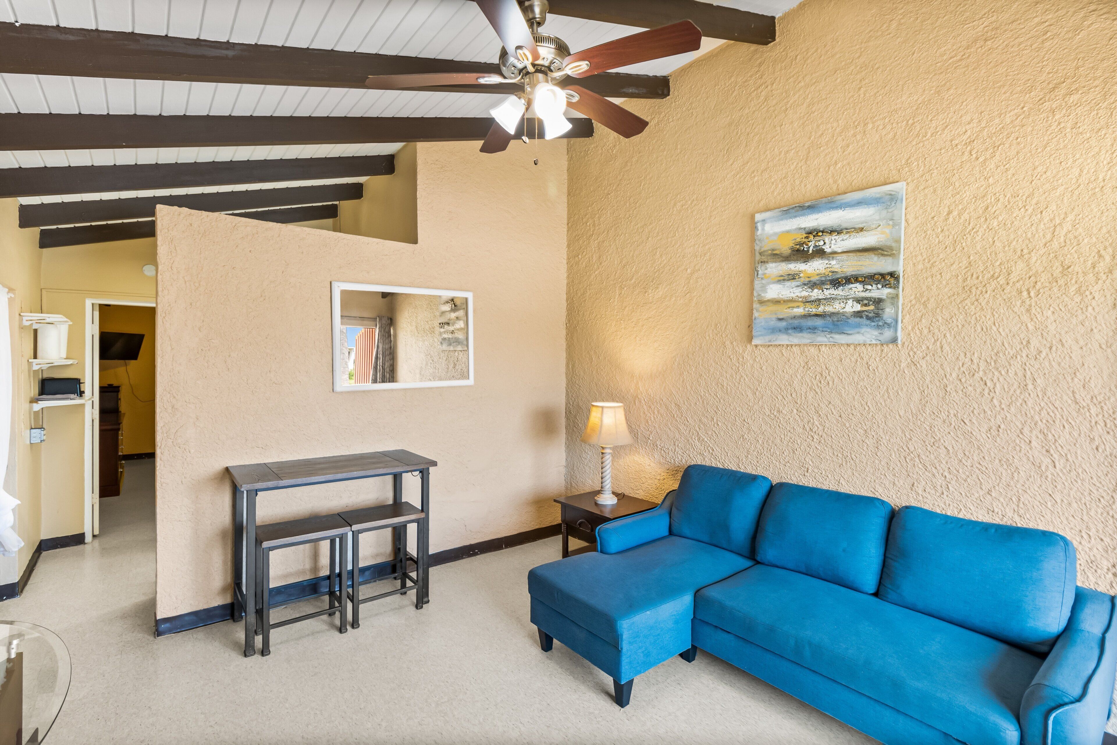 A room with a blue sectional sofa, a small table with benches, a lamp, a ceiling fan, and a painting on the wall.