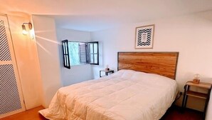 A bedroom with a wooden floor, a bed with a wooden headboard, a nightstand, a wall-mounted light, and a window with shutters.
