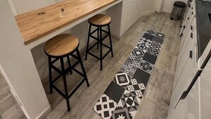 A compact kitchen with a wooden bar counter, two stools, a patterned rug, and a window with a view.